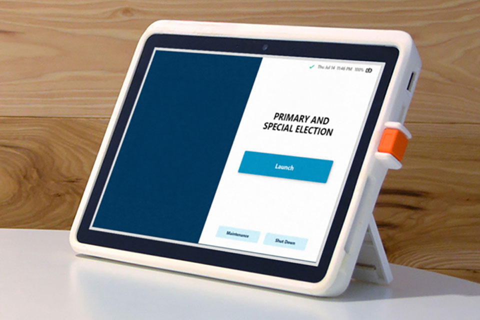 An image shows an Electronic Poll Book on a stand displaying a voting interface for a 'Primary and Special Election.' The screen has a blue section on the left and a white section on the right. In the white section, there are the words 'PRIMARY AND SPECIAL ELECTION' in black text above a blue 'Launch' button. Below the Launch button, there are two smaller buttons labeled 'Maintenance' and 'Shut Down.' The tablet is set against a wooden background, and there is a visible orange clip on the right side of the tablet's case. The screen's top-right corner shows the date and time, 'Thu Jul 14 11:46 PM,' and a battery icon indicating full charge.