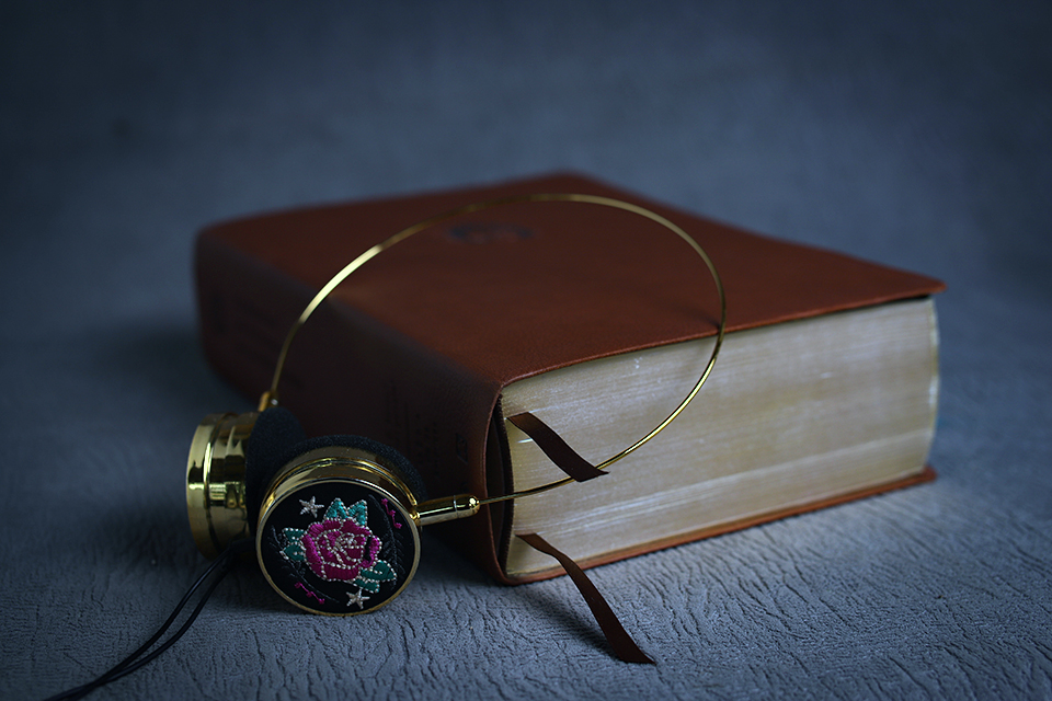 A beautiful pair of ornamental gold headphones with purple embroidered roses on each side adorn a leather bound book that sits closed on a couch with two place markers carfully placed inside.