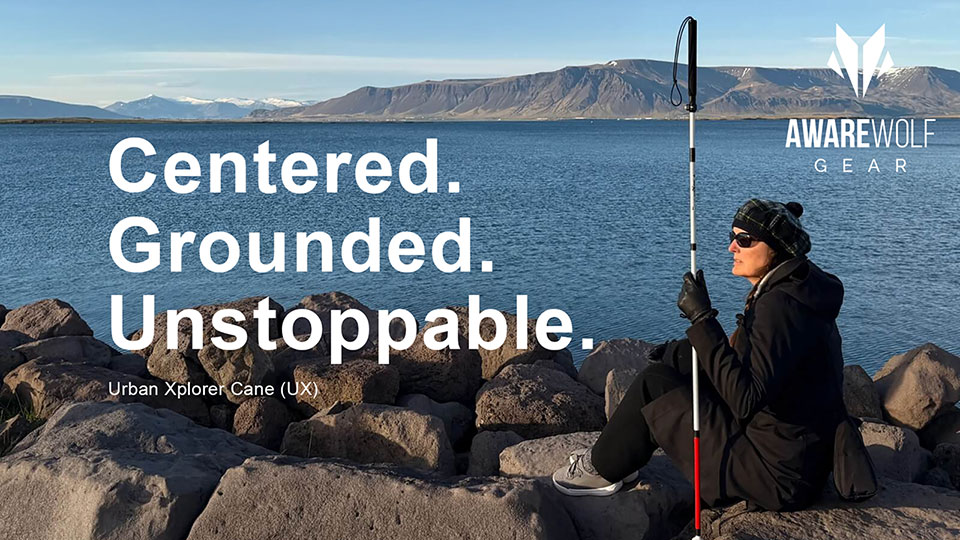 A blind woman dressed in black outdoor clothing, including gloves, sunglasses, and a plaid hat, sits on large rocks at the edge of a serene body of water, holding a white and red Urban Xplorer Cane upright beside her; behind her, calm blue water stretches to distant snow-capped mountains under a clear sky. Large bold text on the left reads: 'Centered. Grounded. Unstoppable.' with smaller text below stating 'Urban Xplorer Cane (UX)'. The Awarewolf Gear logo appears in the upper right corner.