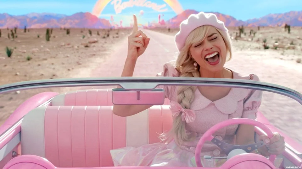 Screen shot from the movie Barbie. Barbie in her car on the road singing with her right index finger pointed into the air.