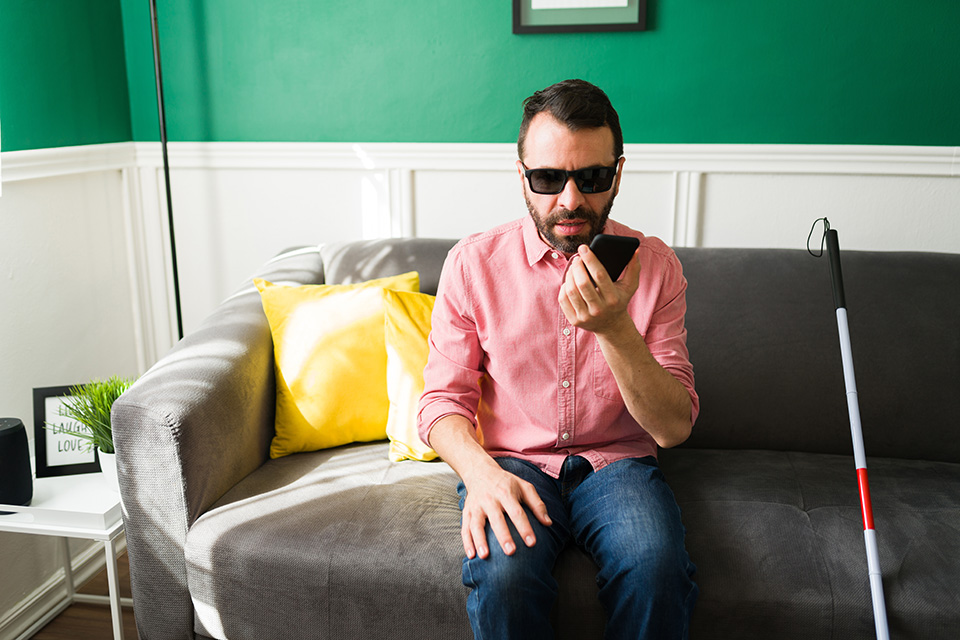 Photo representing a male Tidbits reader. A blind man speaks into his phone which transcribes what he is saying. He sits alone on a grey couch with his white cane next to him on a sunny day.