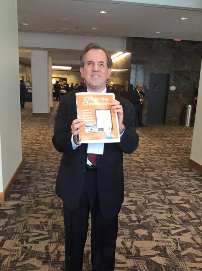 Photo of Larry L. Lewis, Jr. standing in a business suit holding a hardcopy of the book 'iOS In The Classroom.
